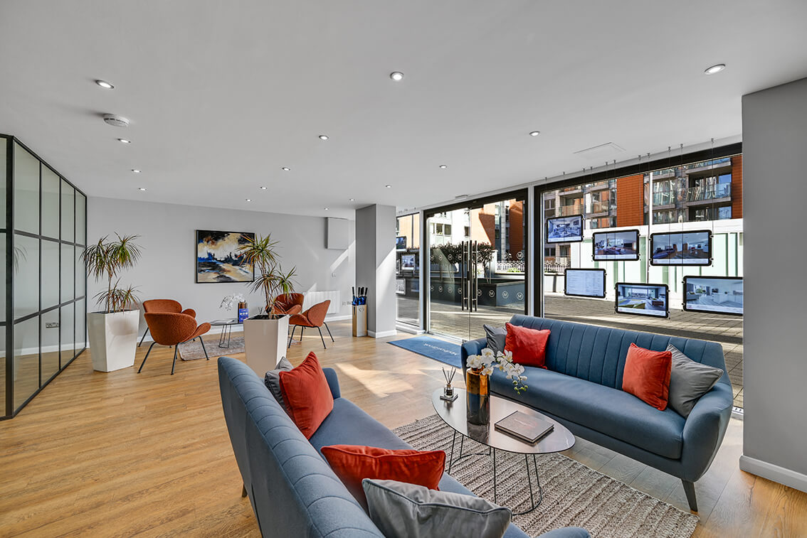 Internal lounge area of an office reception area with 2 blue sofas, a grey table, wooden flooring, four terracotta coloured chairs and a window display.