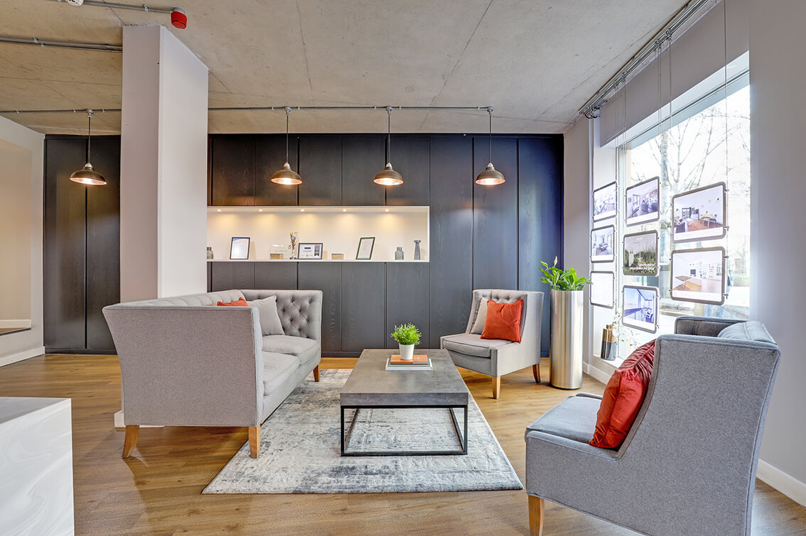 Internal lounge area of an office reception area with a grey sofa, two chairs, a grey table, wooden flooring, grey wall panelling, four vintage style lamps and a window display