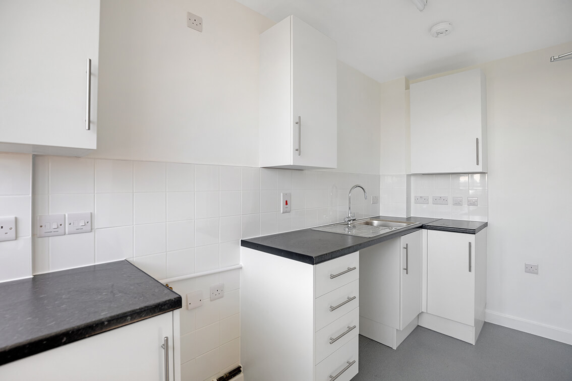 Refurbished kitchen with white kitchen unit, white walls, grey flooring, a sink and two spaces for other white goods.