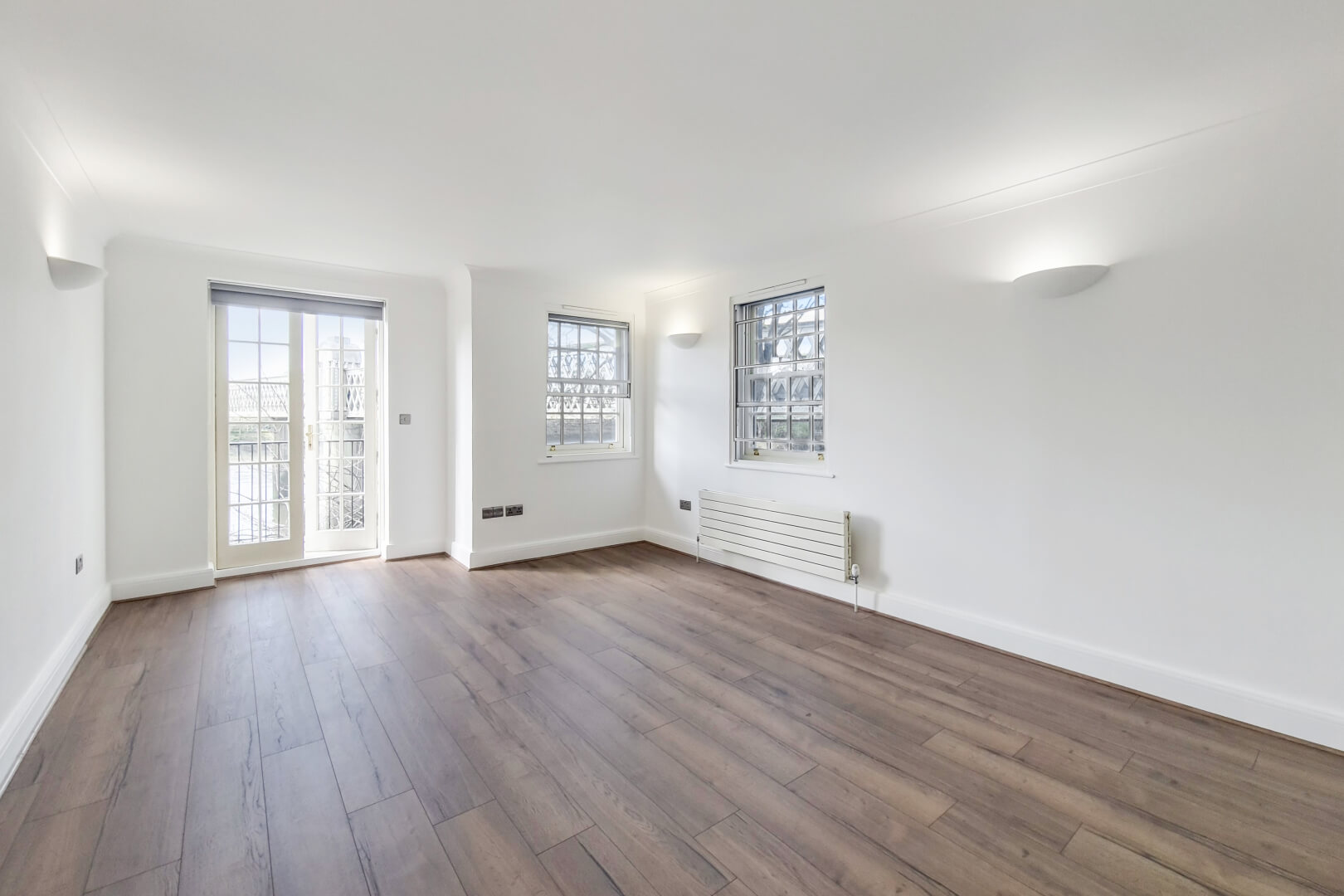 Living area with white walls, wooden floors, uplighter, double aspect windows, a modern radiator and double doors to a balcony