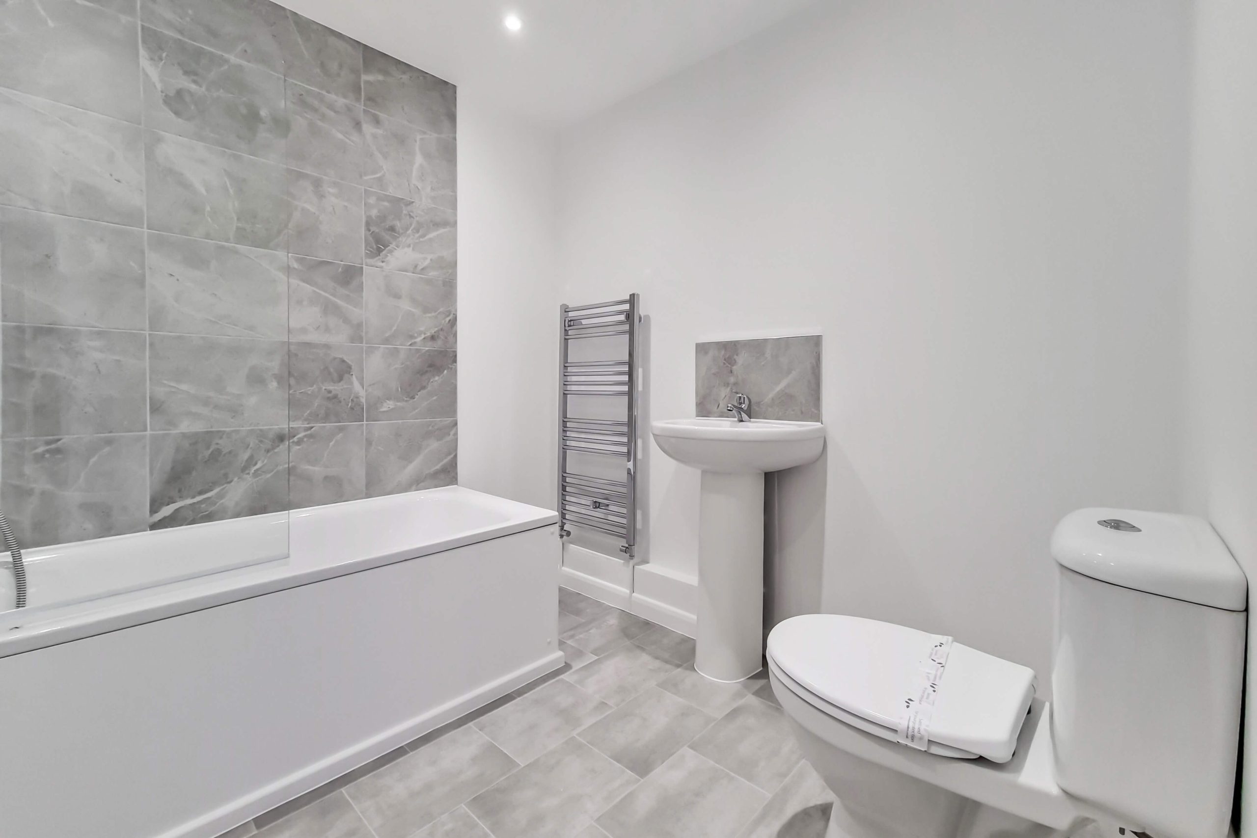 White bathroom suite with WC, handbasin, heated towel rail, bath with overhead shower, white wall and grey tiles