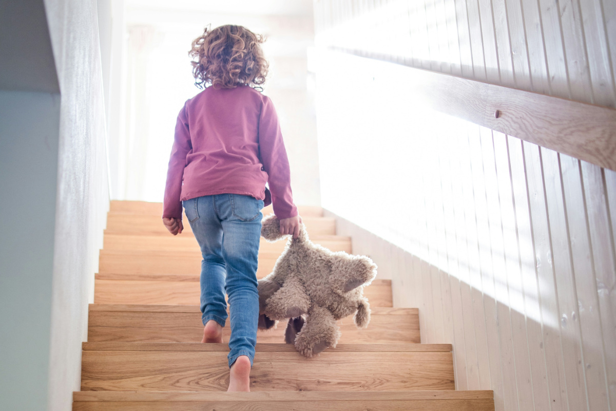 A child walks up wooden stair holding their teddy with light shining from the upstairs window